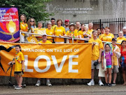 A collection of adults and children in yellow shirts stand around a yellow banner inscribed with the words "Side with Love."