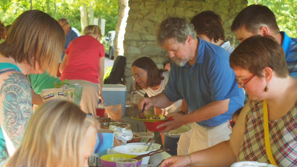 A group of adults, children, and youth, crown around a picnic table for a potluck lunch.