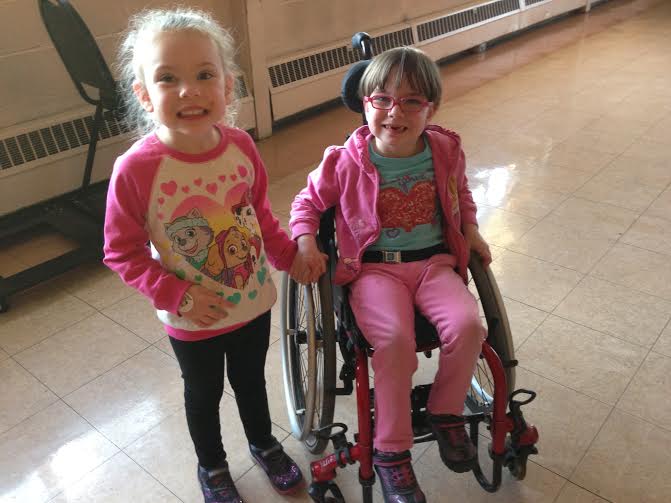 One child in a wheelchair, and another child who is standing, hold hands.  They are both wearing pink hearts in celebration of Valentine's Day.