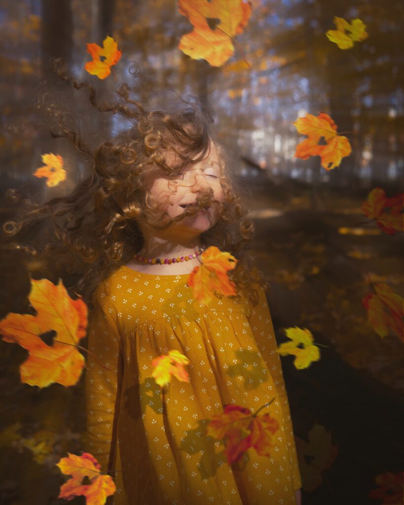 A girl with curly hair is standing in a windy forest.  All around her leaves fly.  Some leaves contain cutouts in the shape of a heart.
