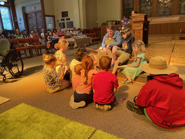 A larger white woman with short hair sits on the steps of a small stage with a cardboard box.  She is surrounded by 9 children, who are all listening very intently to her.  They are all bathed in beams of sunlight.