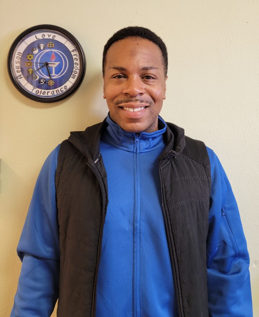 A brown skinned man with close cropped hair and a pencil thin mustache shares a warm smile.  He is wearing fleece under a black vest as he stands in front of a cream colored wall.  To the left of his head is an interfaith clock.