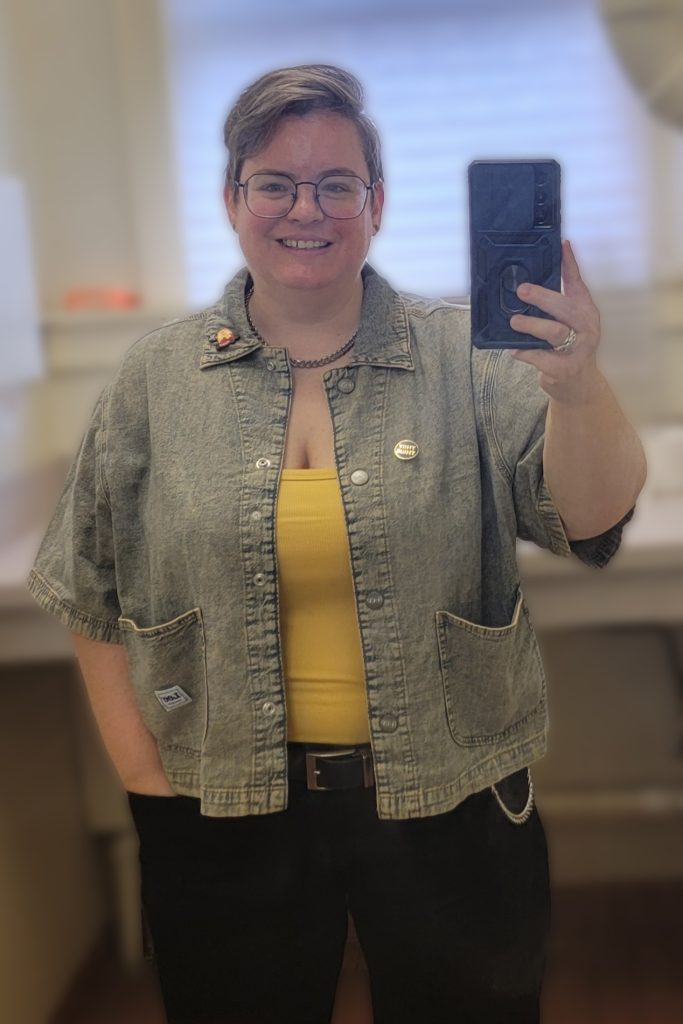 A selfie of Mx Meredith, August 2024. They're wearing glasses, black jeans, a yellow tank and a short sleeve, acid washed jean jacket. They have short brown hair that is shaved on the sides and long on top. They are adorned with several pieces of jewelry, including a pronoun pin, and a chain wallet.