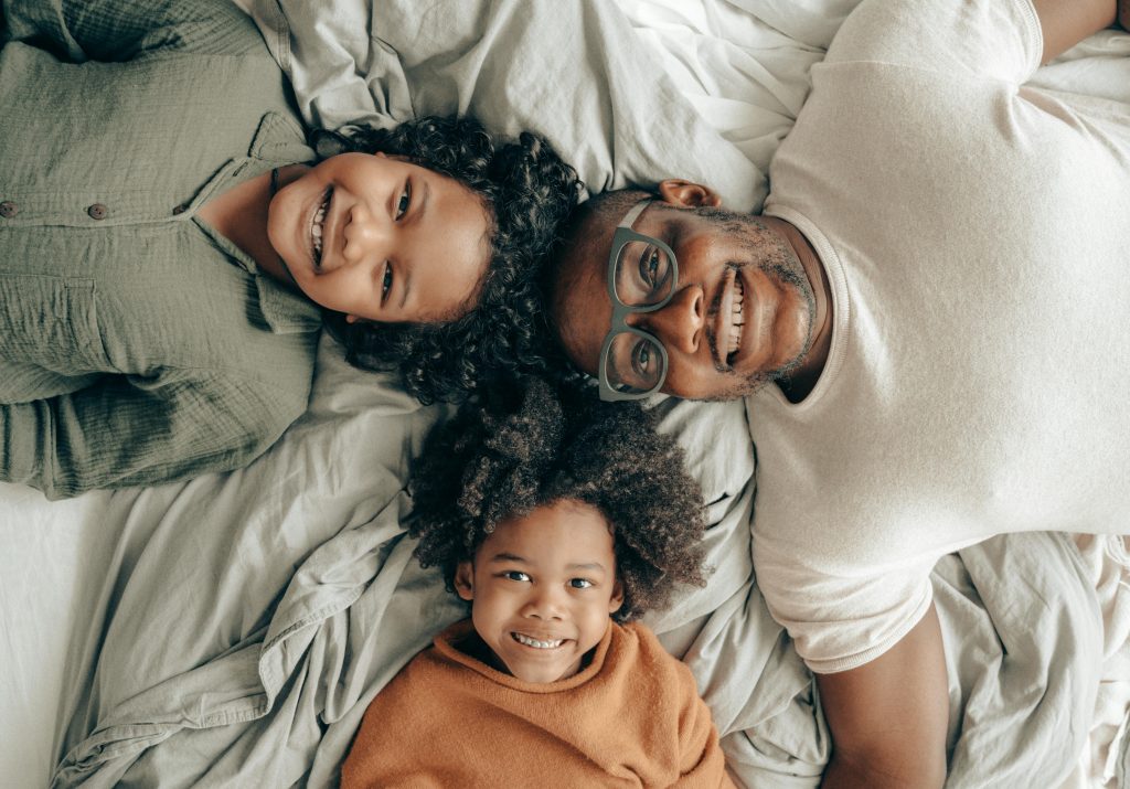 A black family - dad and two kids - lie on a bed with their heads together, smiling up at the camera.