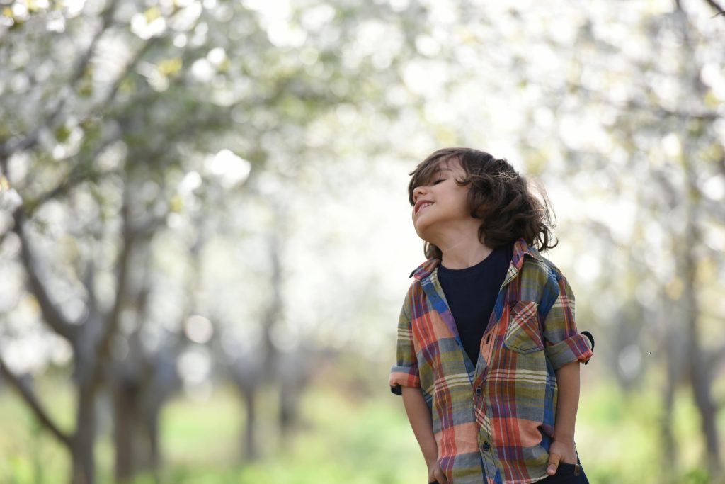 A young child of about 5 with shoulder length hair stands in nature and smiles up at the sky.